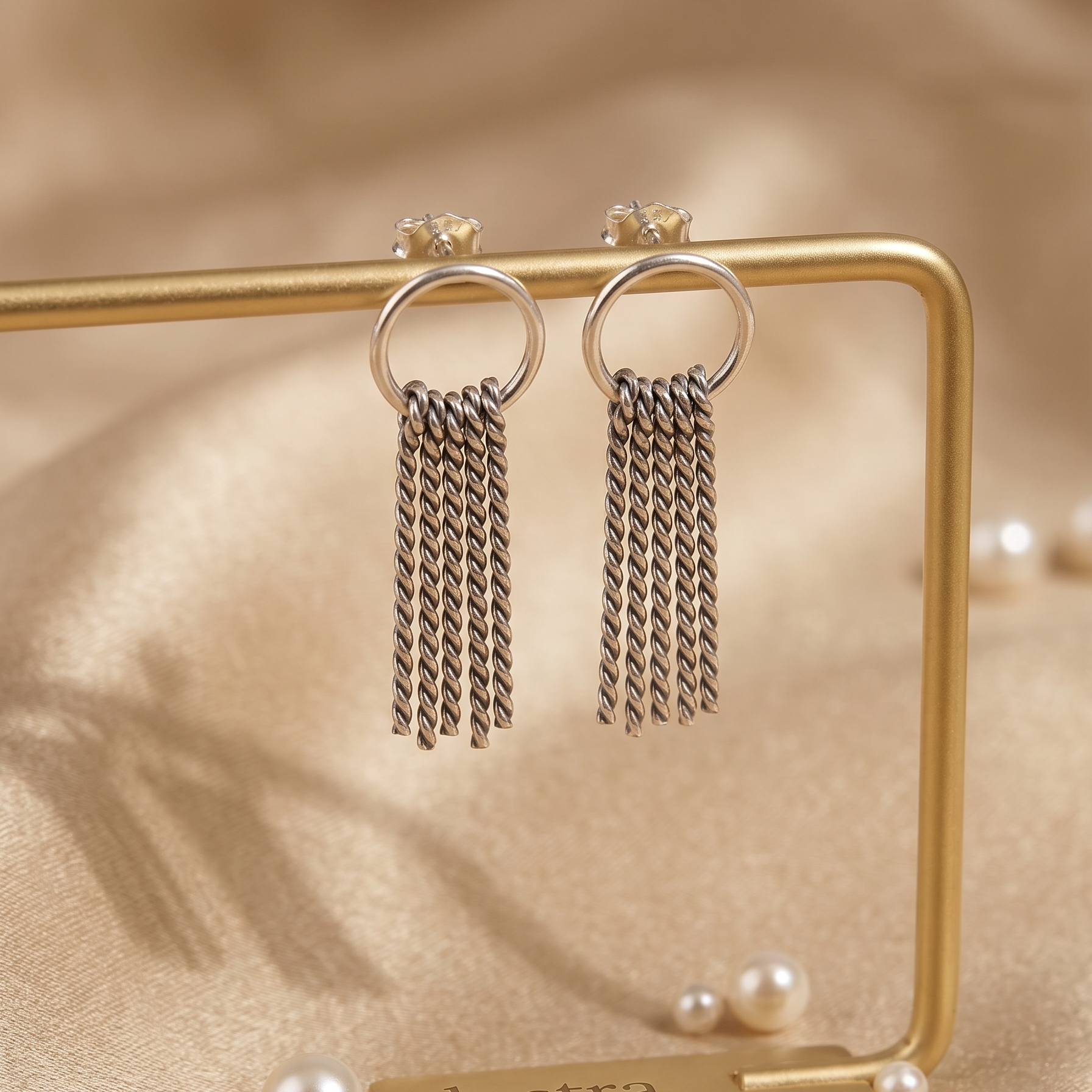Pair of silver chain earrings on a gold stand with 'lustra' branding, set against a beige fabric background.