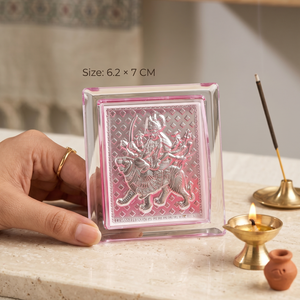 Small decorative item with a religious design held by a hand on a surface with incense and a lamp.