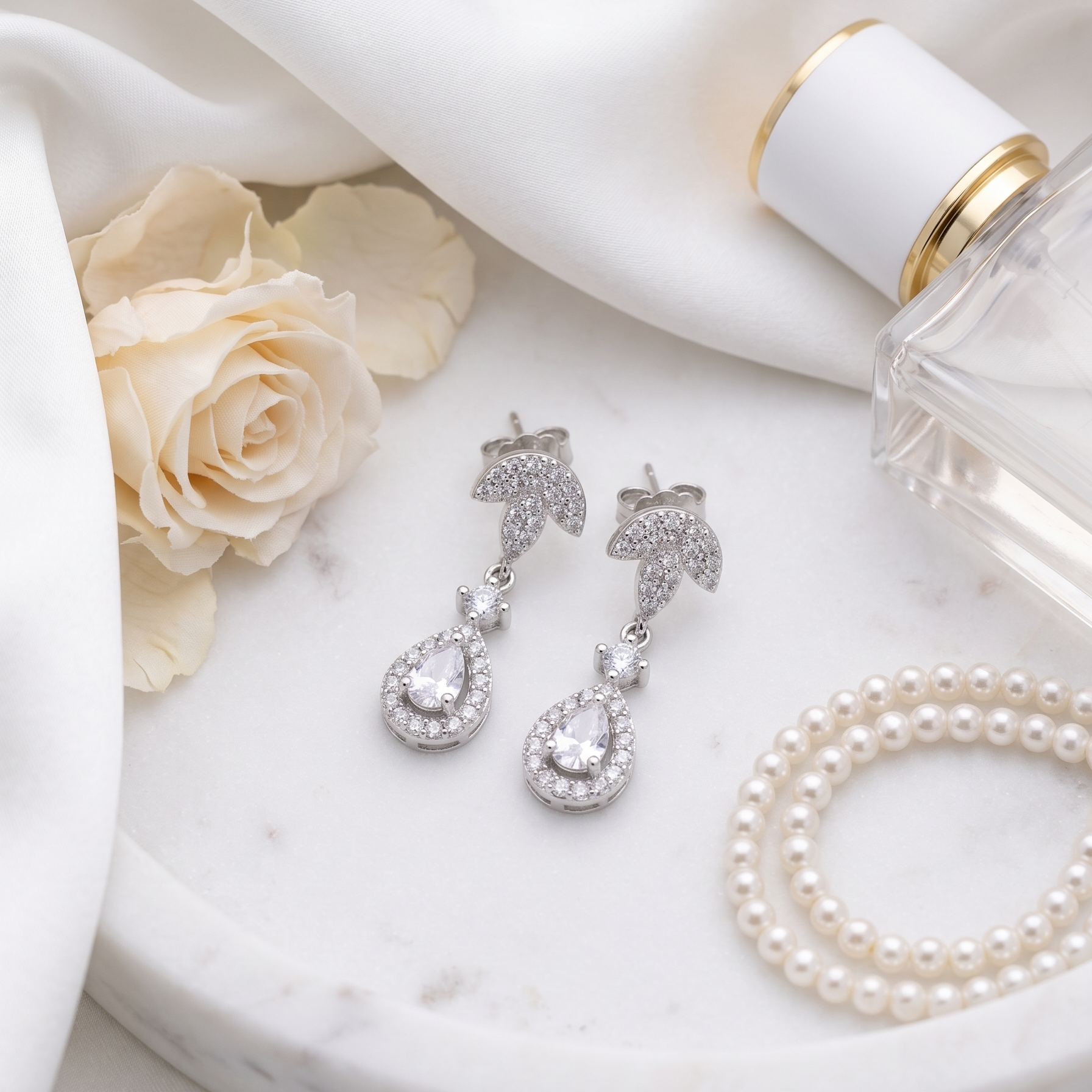 Silver earrings with pearls on a marble surface with a white rose and perfume bottle.