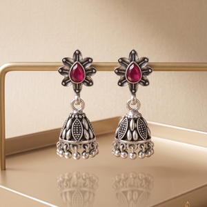 Pair of silver jhumka earrings with red gemstones on a reflective surface.