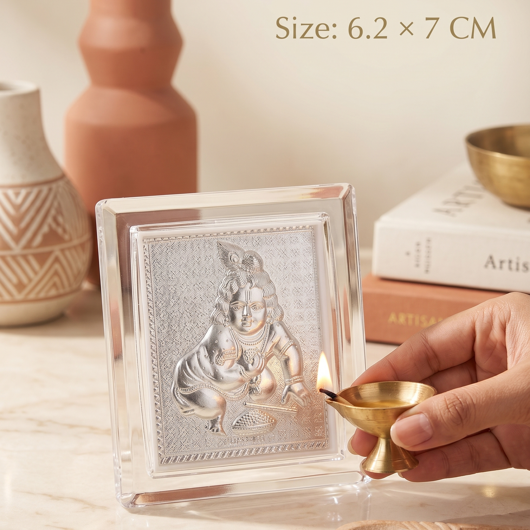 Small silver deity statue in a clear crystal frame with a hand holding a lit candle on a wooden surface.